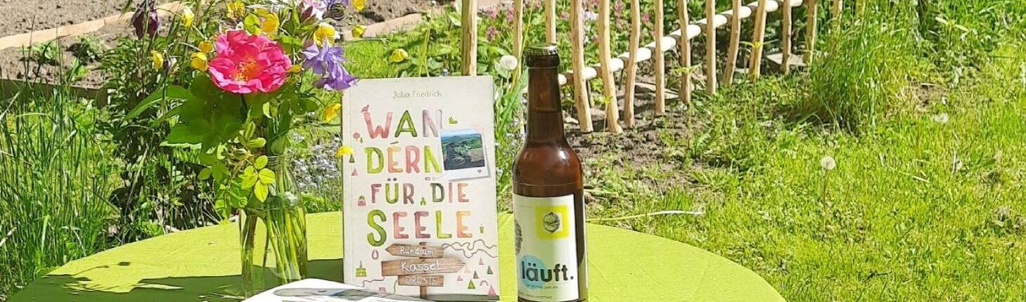 Der Wanderführer Wandern für die Seele - Rund um Kassel zwischen einem Blumenstrauß und einem regionalen Bier auf einem Tisch mit früner Tischdecke präsentiert.