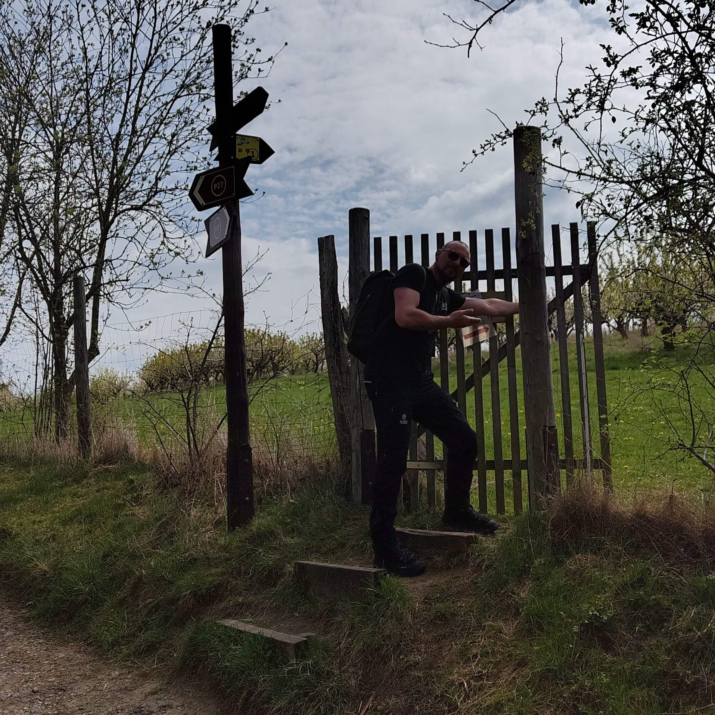 Person öffnet ein Törchen in einem Zaun neben einem Wegweiser.